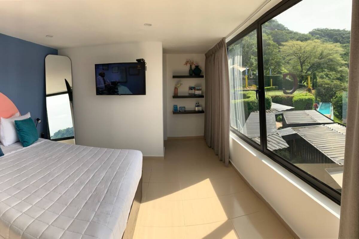 A bedroom with a bed, a chair, a TV, and a large window with a view.