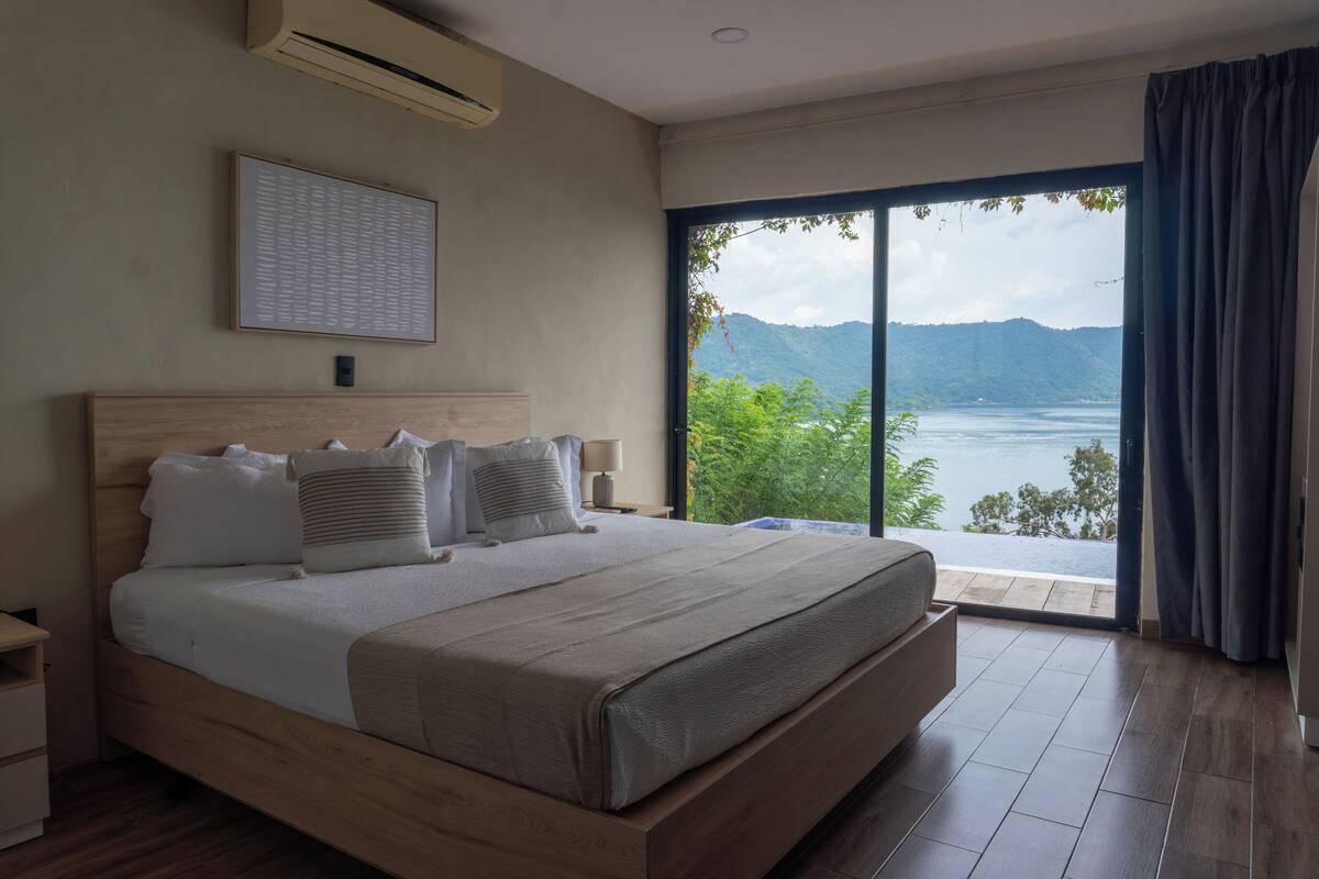 A bedroom with a large bed, a nightstand, a wall-mounted air conditioner, and a window with a view of a lake and mountains.