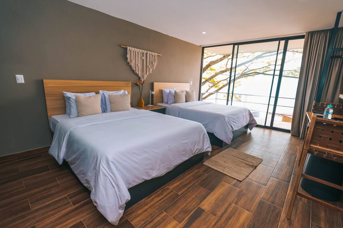 A modern hotel room with two beds, wooden flooring, and large windows offering a view of trees.