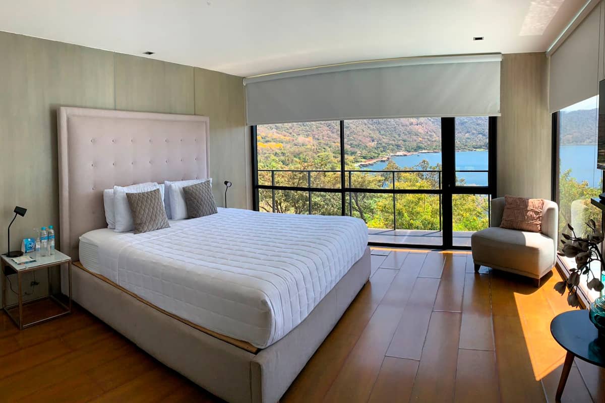 A bedroom with a large bed, a nightstand, a chair, and a view of a lake and mountains.