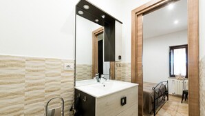 Een badkamer met een grote spiegel, een wastafel en een douche. In de aangrenzende kamer staat een bed en is er een raam.