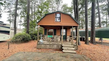 Cozy cabin with a front porch and a small window above the door, set amidst a wooded area.
