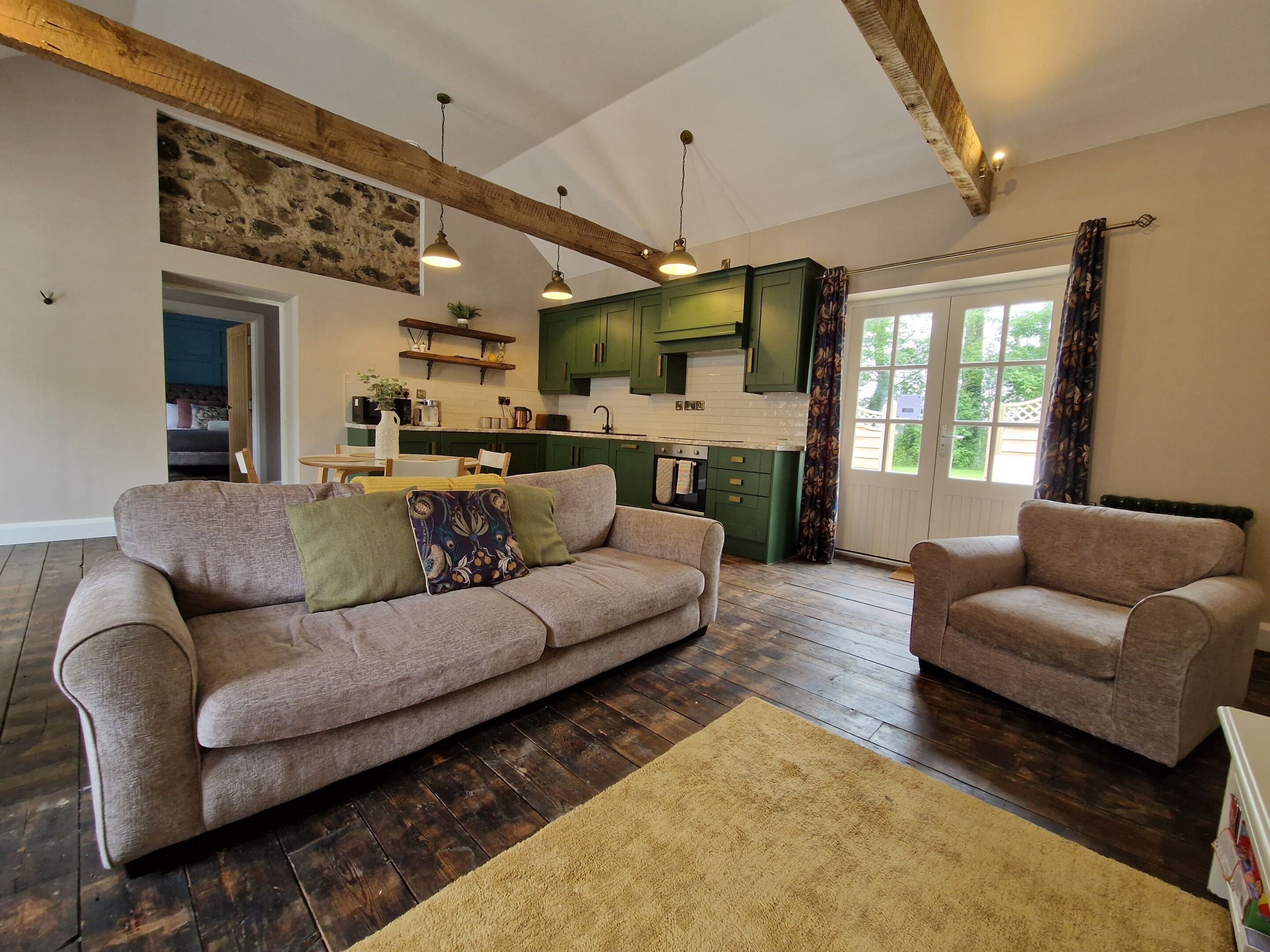 Una sala de estar con sofá y sillón, suelo de madera y una cocina al fondo.