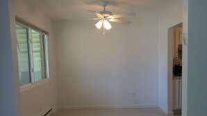 A room with a ceiling fan, a window with blinds, and a kitchen area in the background. - Summer Rental - Great Location - West End Section of Long Branch - Walk to beach (Long Branch)