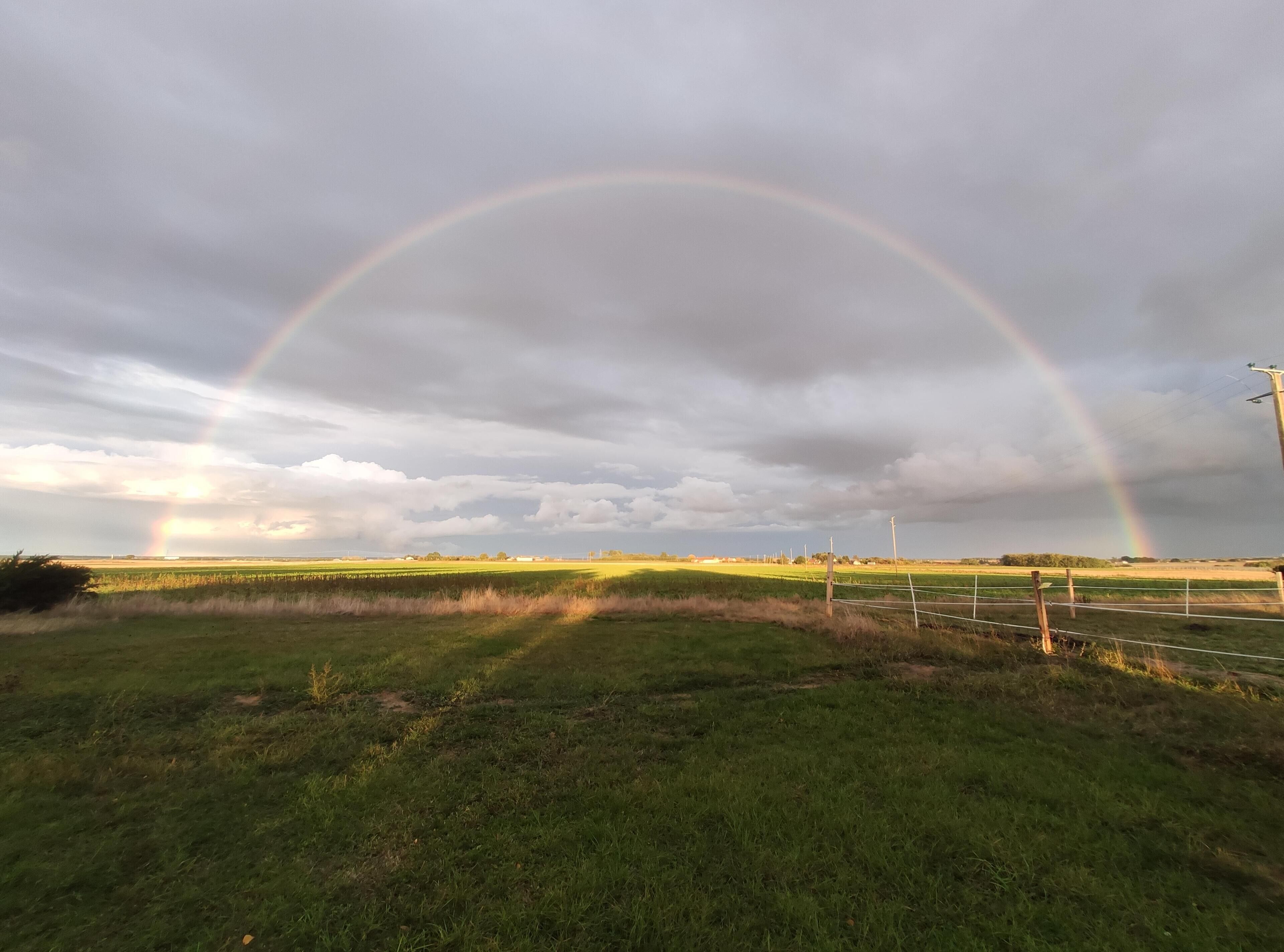 A rural landscape with a rainbow over a field, a fence, and a cloudy sky.