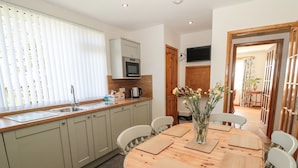 A modern kitchen with a dining area, featuring a round wooden table, light-colored chairs, a built-in microwave, and a large window with blinds. - Rose Bank (Belford)