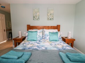 Un dormitorio con cabecera de madera, colcha estampada, dos almohadas a juego, una manta gris y dos mesitas de noche con lámparas blancas.