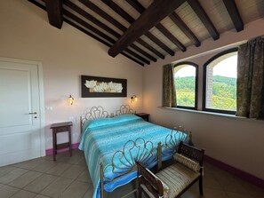 Un dormitorio con cama, una banca, una mesita de noche, una lámpara de pared y una ventana con vistas a la vegetación.
