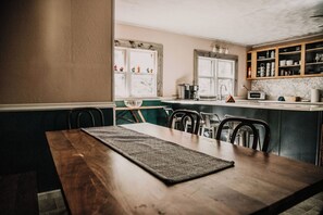 A dining area with a wooden table and chairs, a kitchen in the background with a window, and a shelf with kitchenware.