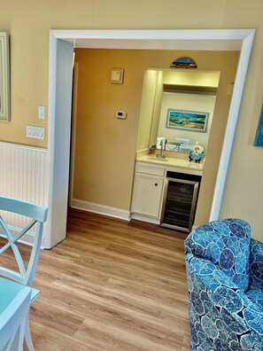 Une pièce avec un plancher en bois, une chaise blanche, un canapé bleu à motifs, un coin cuisine avec un évier et une cuisinière, et une porte donnant sur une autre pièce.