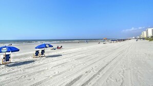 A wide sandy beach with several blue umbrellas and lounge chairs, a clear sky, and buildings in the background. - Oceanfront 3 BR 2.5 bath Tilgham Beach & Racquet Club # 307. 2 pools, Tennis (North Myrtle Beach)