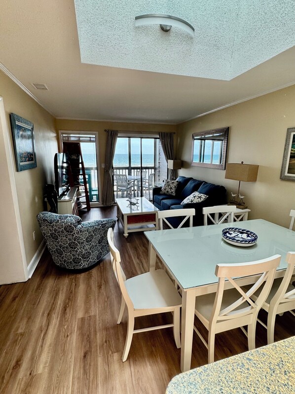 Een ruime woonkamer met balkon en uitzicht op zee, een eettafel met stoelen, een bank en een lamp.