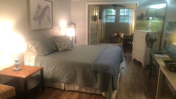 A bedroom with a bed, bedside tables, a dresser, a desk, a chair, a fan, and a window with a view of the outdoors. - Charming Bungalow suite in Peaceful Sea Palms (St. Simons Island)