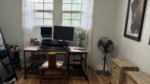 A room with a wooden desk, two computer monitors, a chair, a standing fan, and a framed picture on the wall. - Charming Bungalow suite in Peaceful Sea Palms (St. Simons Island)
