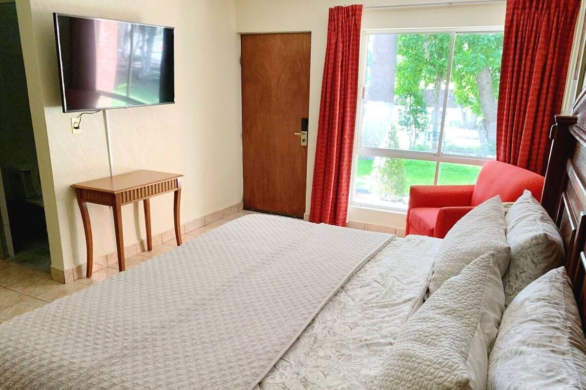 A bedroom with a large bed, a TV mounted on the wall, a wooden nightstand, and a window with curtains.