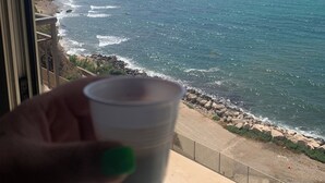 A hand holding a cup near a window overlooking a beach with waves and mountains in the distance. - Window to the Sea by Alexander 2p (Balestrate)