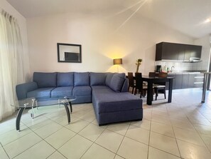 A modern living room with a sectional sofa, a glass coffee table, a dining area with a table and chairs, and a kitchen area with cabinets and a stove. - Jade Beach A6 | Walk to Beach, Dining & Shops (Playas del Coco)