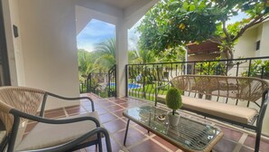 Un balcón con mobiliario de mimbre, una mesa de vidrio y vistas a una piscina y follaje.