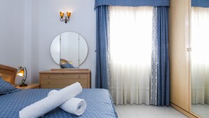 Un lit bien fait, avec une serviette roulée, une lampe de chevet, un miroir rond, une commode en bois et une armoire avec un miroir.