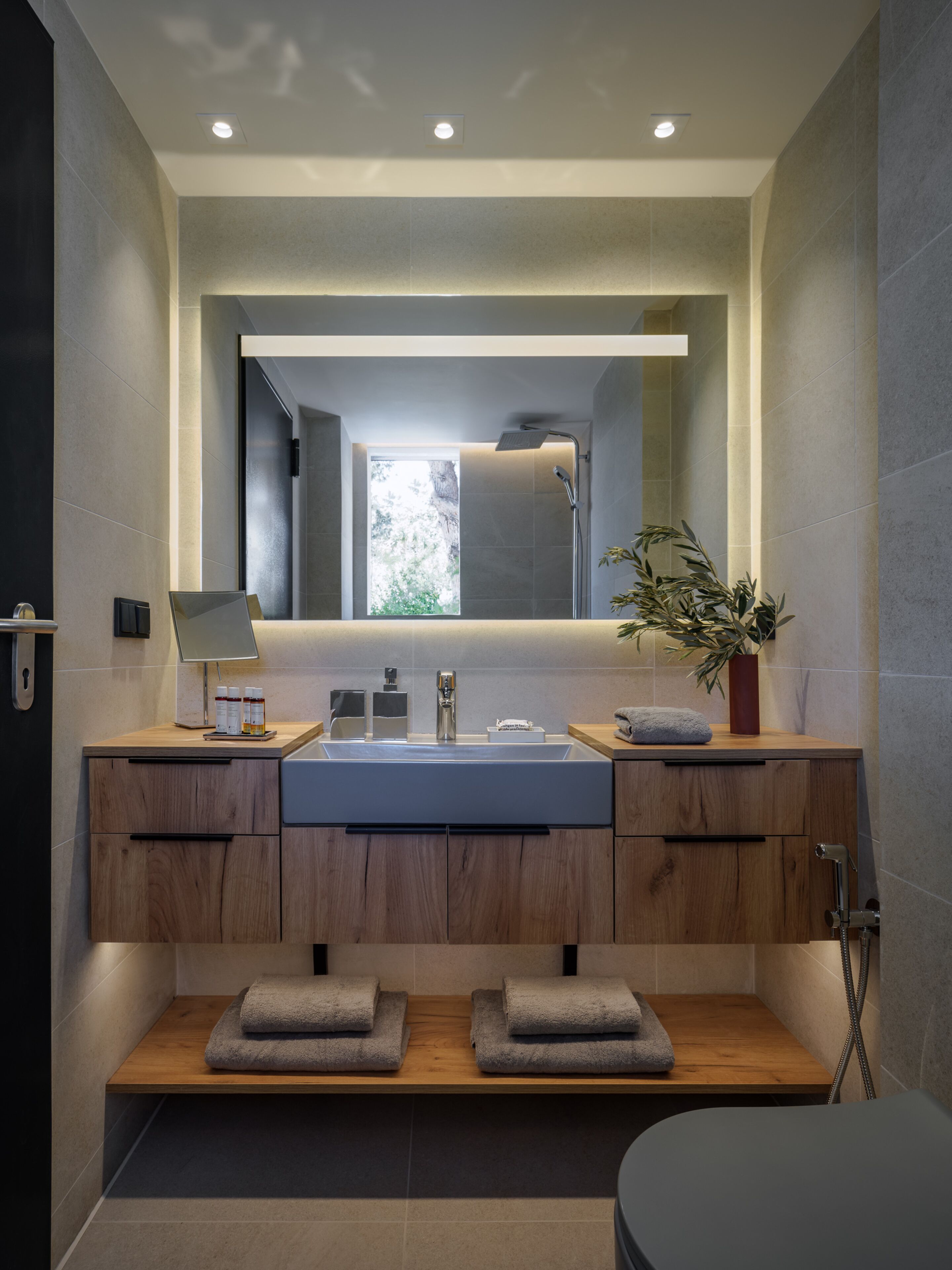 Ein modernes Badezimmer mit großem Spiegel, zwei Waschbecken, Holzschränken und einer Bank mit Handtüchern.