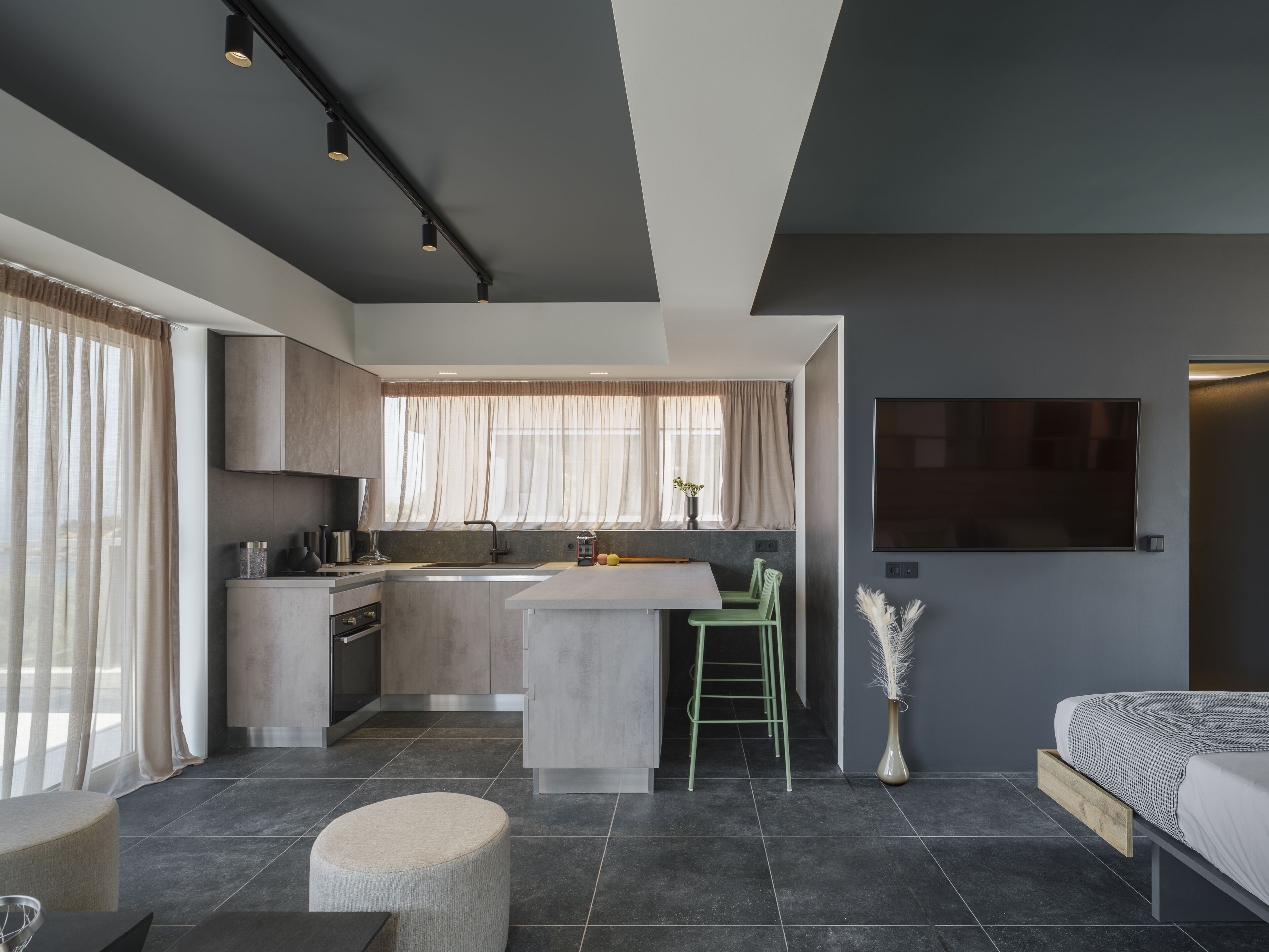 A modern kitchen with a central island, a dining area with a green chair, a bed with a TV, and a large window with sheer curtains.