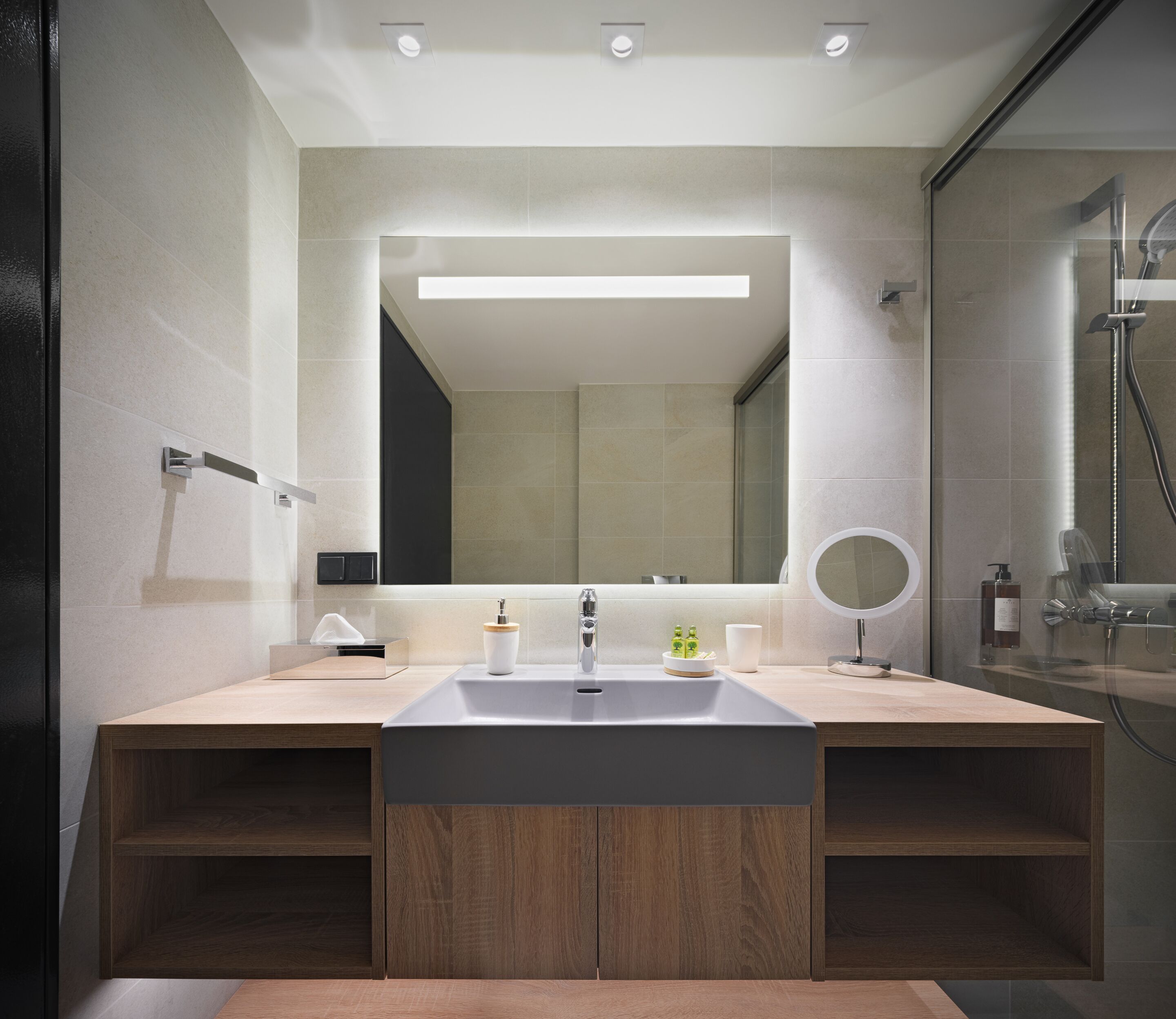 A modern bathroom with a large mirror, two sinks, a shower, and a wooden vanity.