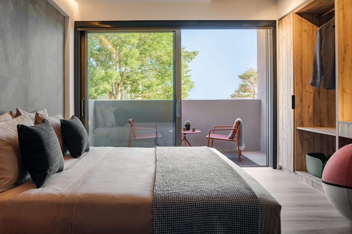 Une chambre moderne avec un grand lit, une porte-fenêtre coulissante donnant sur un balcon et une armoire en bois.