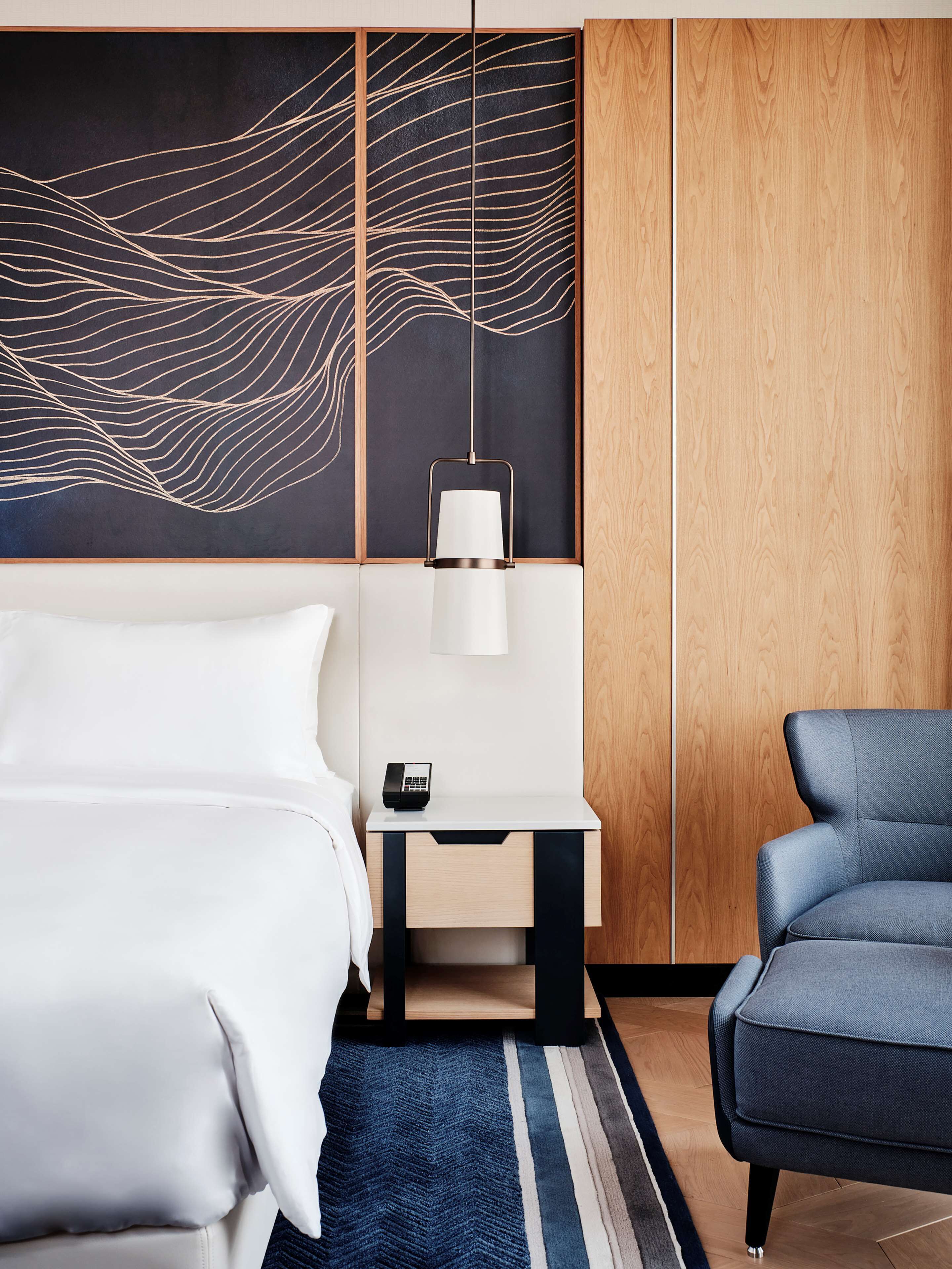 A modern hotel room with a bed, a desk with a lamp, a chair, and a decorative wall panel.
