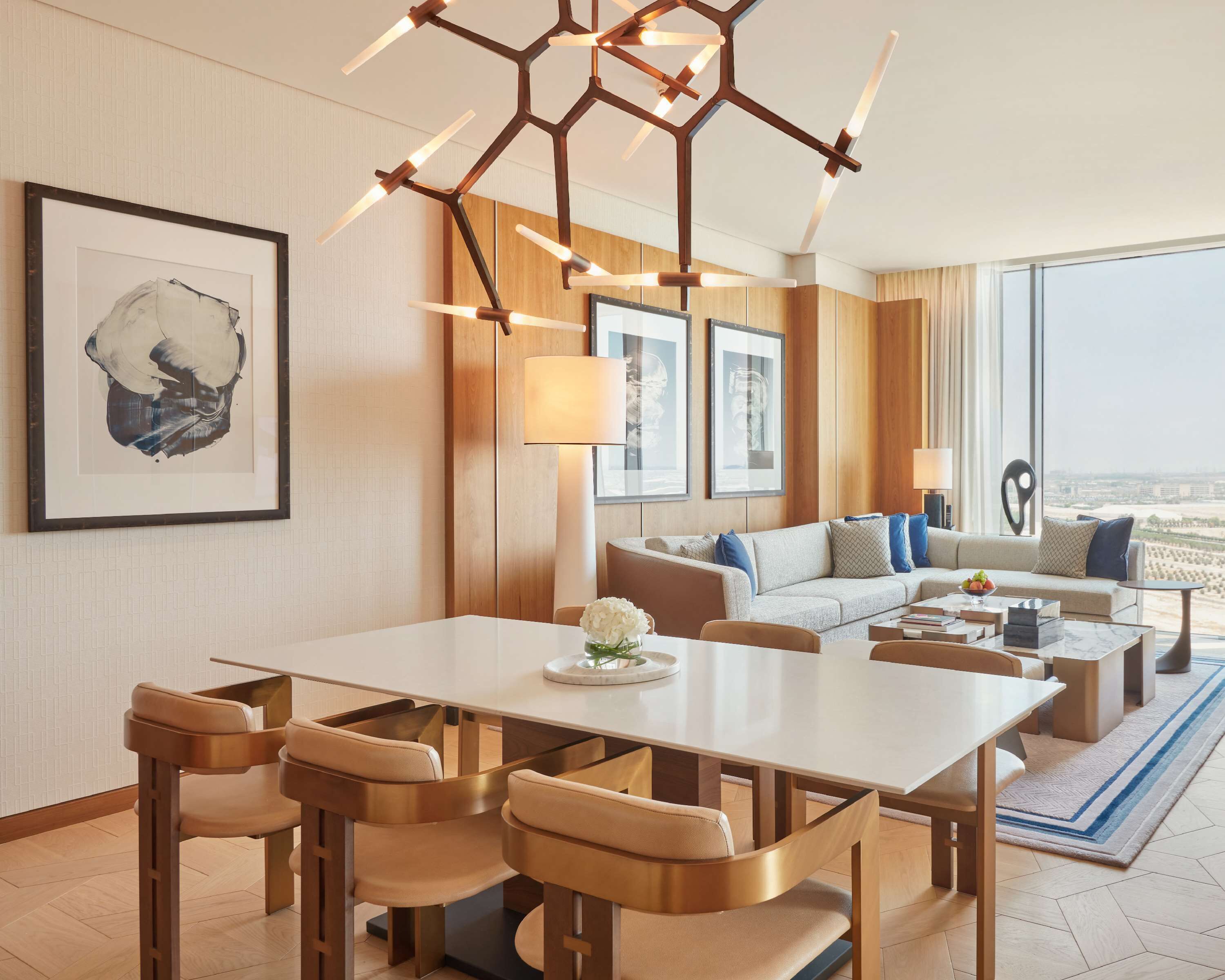 A modern hotel room with a dining area, a sofa set, and large windows offering a city view.