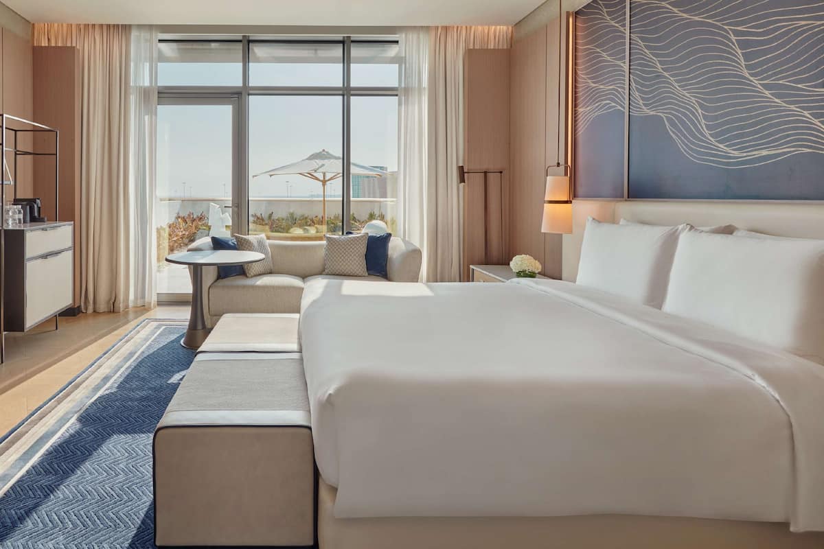 A hotel room with a large bed, a desk, a chair, a small table with a lamp, and a view of the city through large windows.