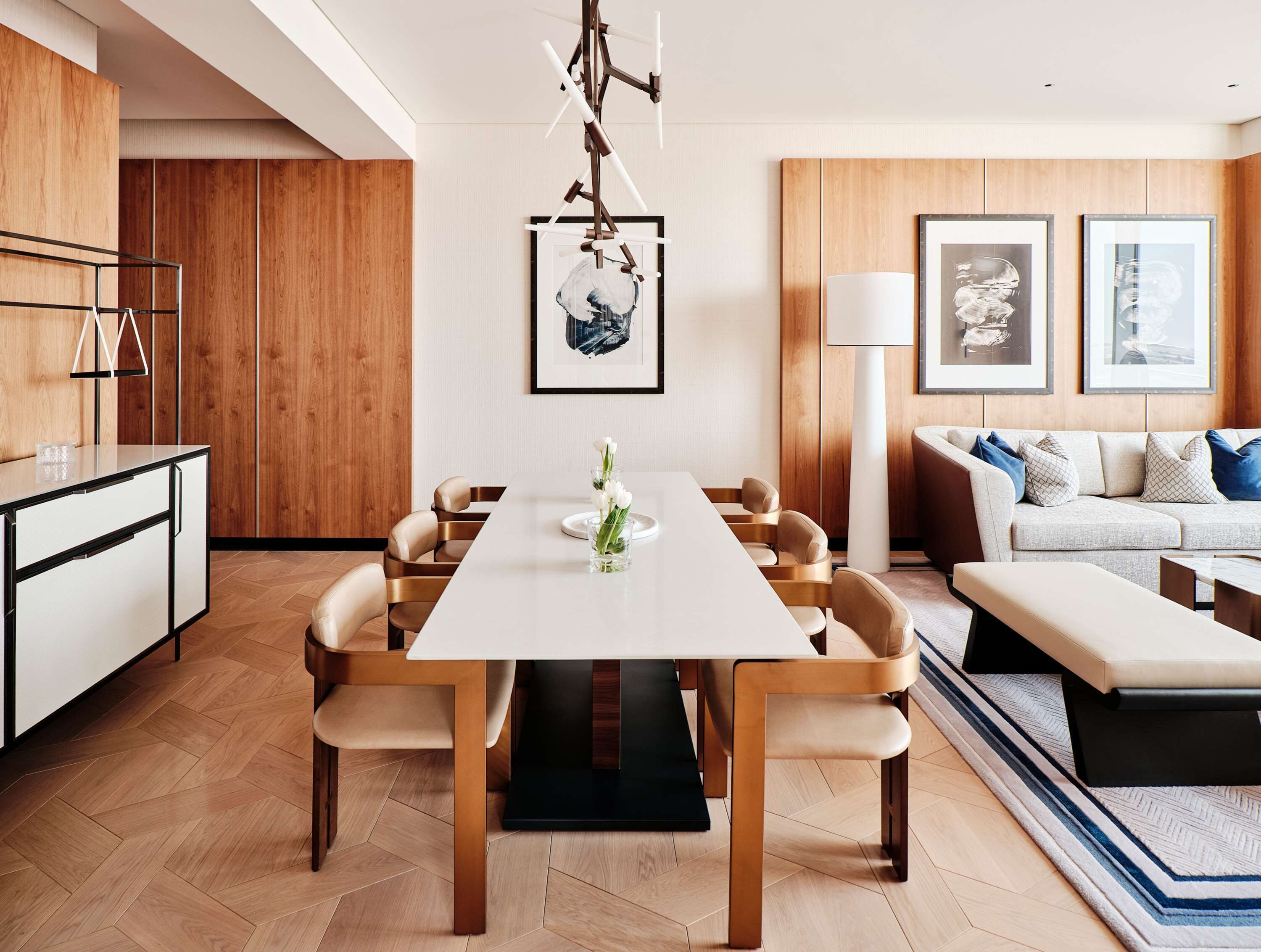 A modern dining area with a white table, beige chairs, and a wooden floor. There is a sofa and a lamp in the background.