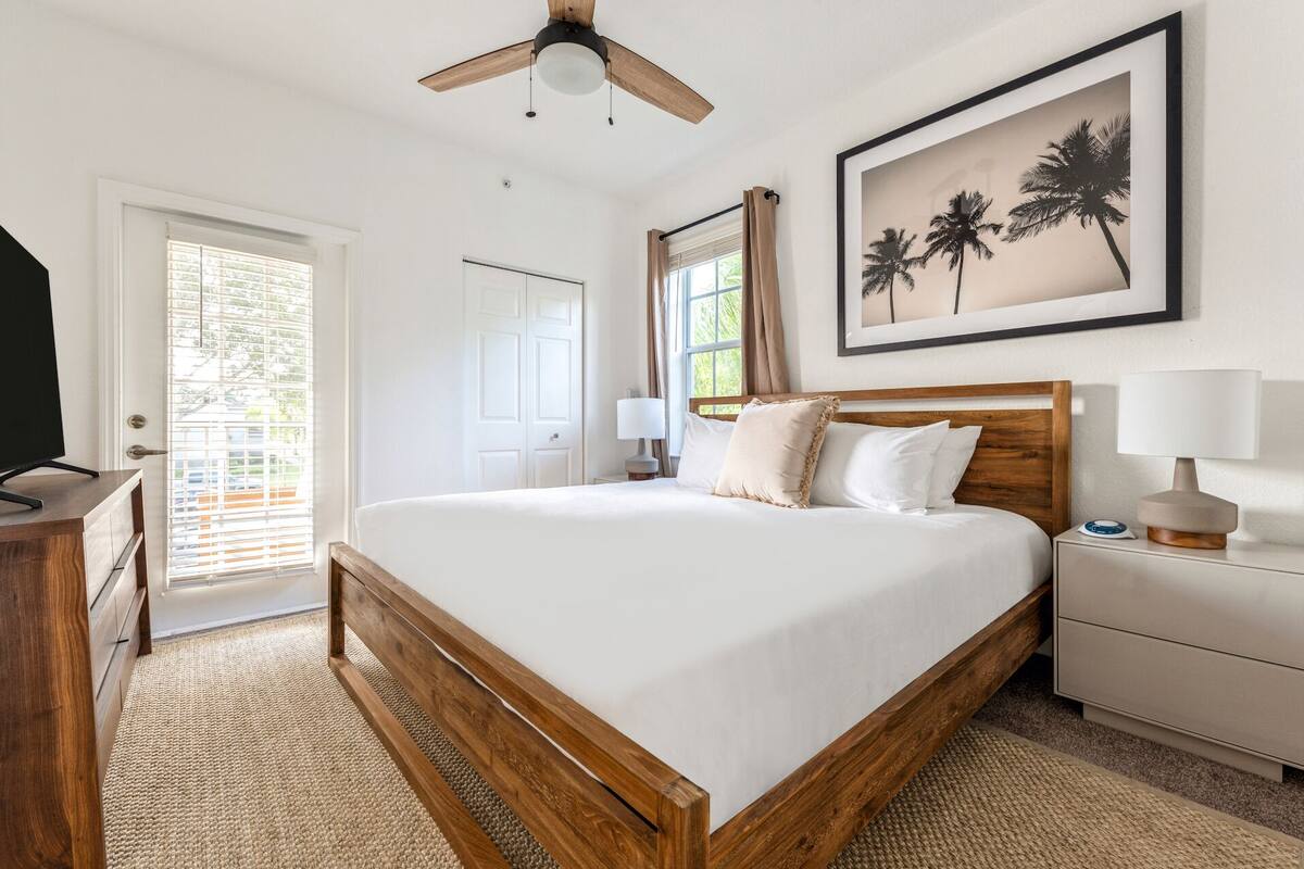 A bedroom with a wooden bed frame, white bedding, a ceiling fan, a framed picture of palm trees, a bedside table with a lamp, and a door with blinds.