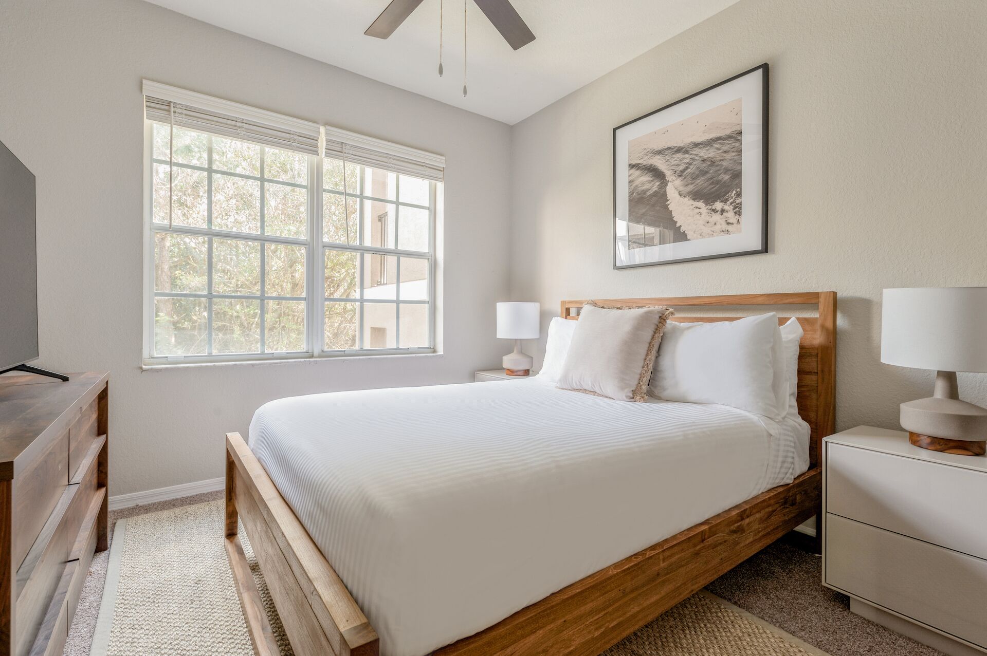 A bedroom with a large window, a bed with white bedding, a wooden headboard, two bedside tables with lamps, and a framed picture on the wall.