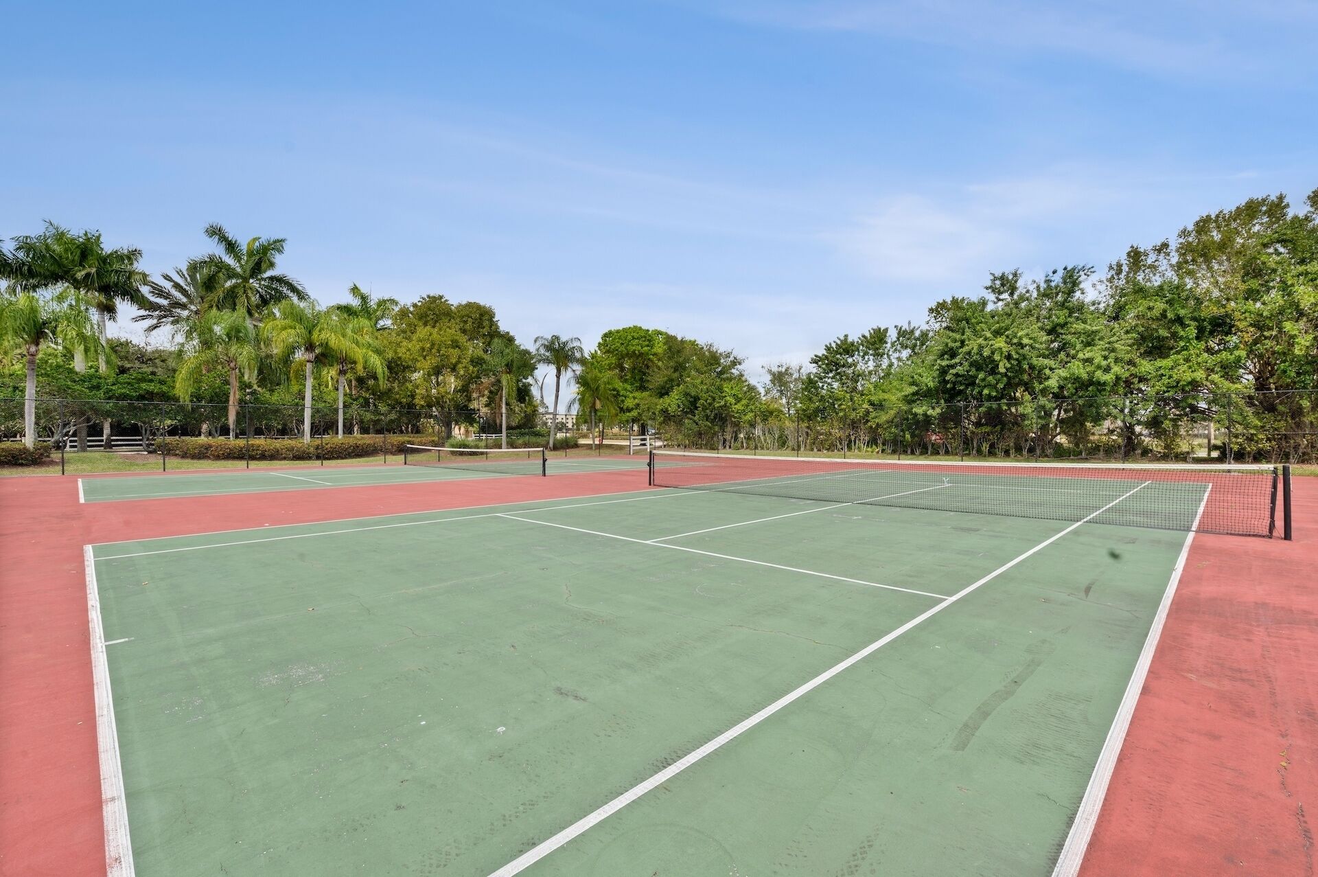 An outdoor tennis court with multiple courts, surrounded by trees and a clear sky.