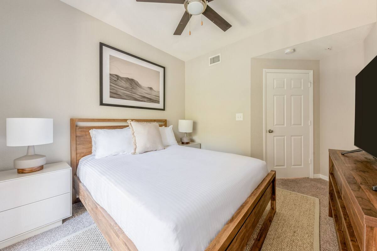 A neatly arranged bedroom with a bed, bedside tables, a ceiling fan, a door, and a television.