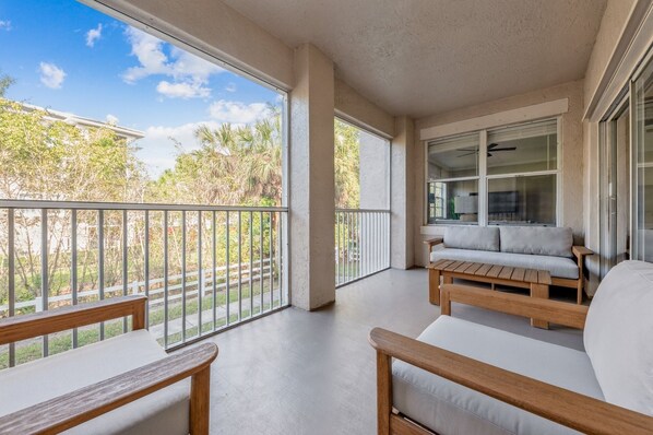 A spacious balcony with a wooden bench and a sofa, large windows, and a view of greenery and a house.