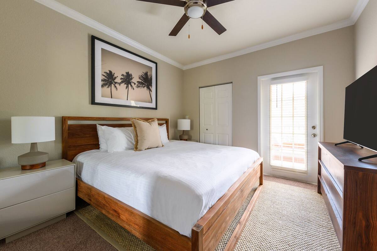 A bedroom with a wooden bed, a ceiling fan, a framed picture of palm trees, a white door, and a television on a wooden dresser.