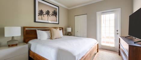 A bedroom with a wooden bed, a ceiling fan, a framed picture of palm trees, a white door, and a television on a wooden dresser.