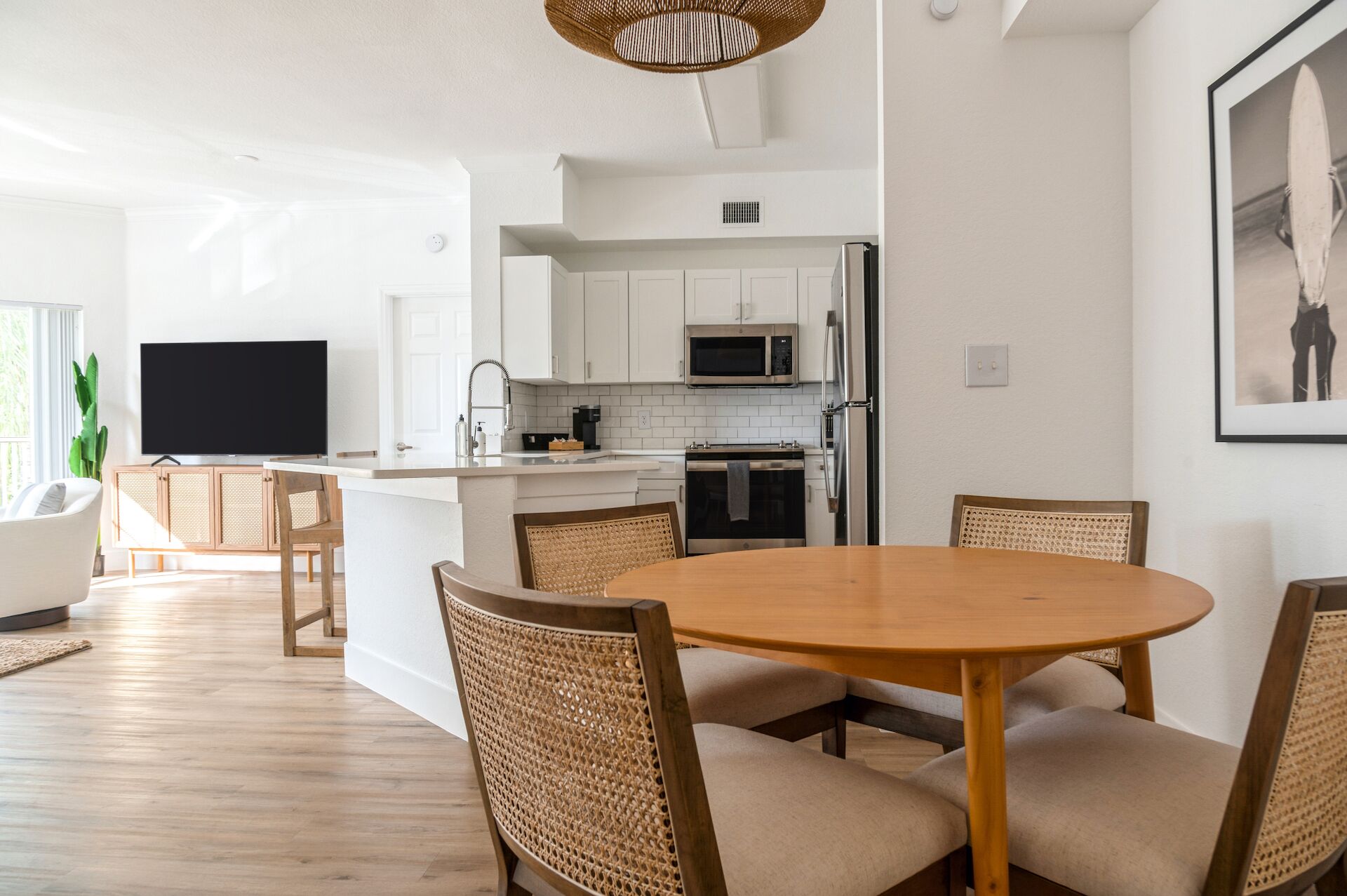 A modern kitchen with white cabinets, a microwave, and a refrigerator. There is a dining area with a round wooden table and wicker chairs.