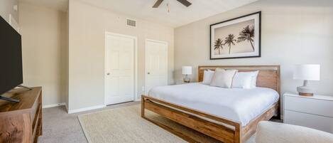 A bedroom with a wooden bed, white bedding, a ceiling fan, two white doors, a framed picture of palm trees, and a bedside table with a lamp.