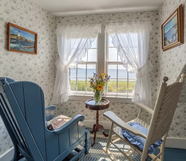 A room with a view of the sea, featuring a blue rocking chair, a small table with a vase of flowers, and a wicker chair.