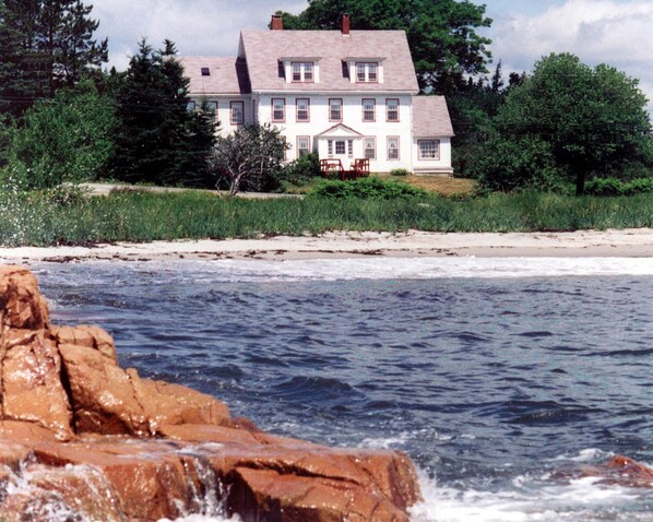Una casa grande de dos pisos con fachada simétrica, un porche delantero y un garaje visible. La casa está ubicada en una playa arenosa con un litoral rocoso en primer plano.