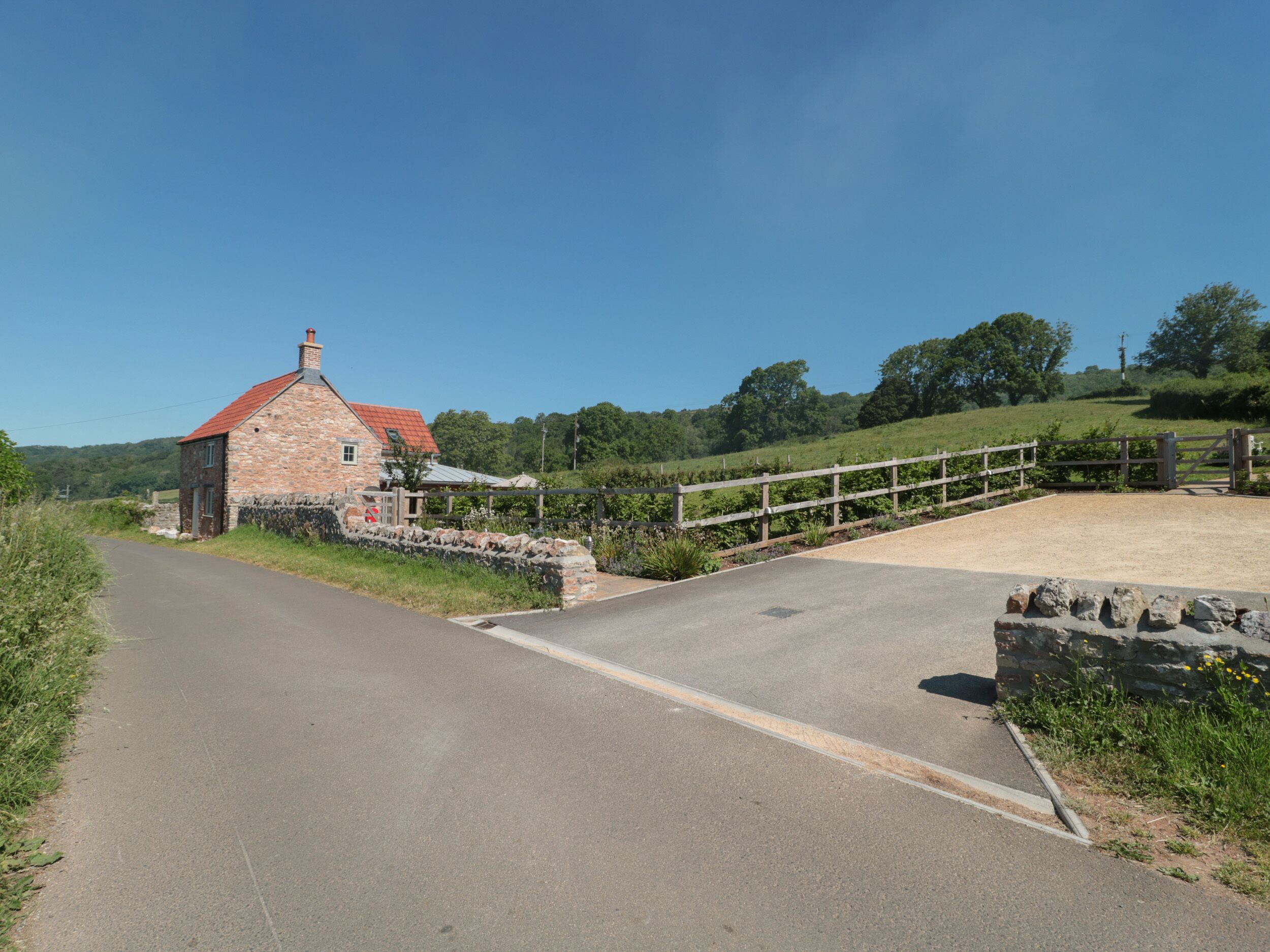 A rural road leading to a stone building with a red roof, surrounded by greenery and a wooden fence.