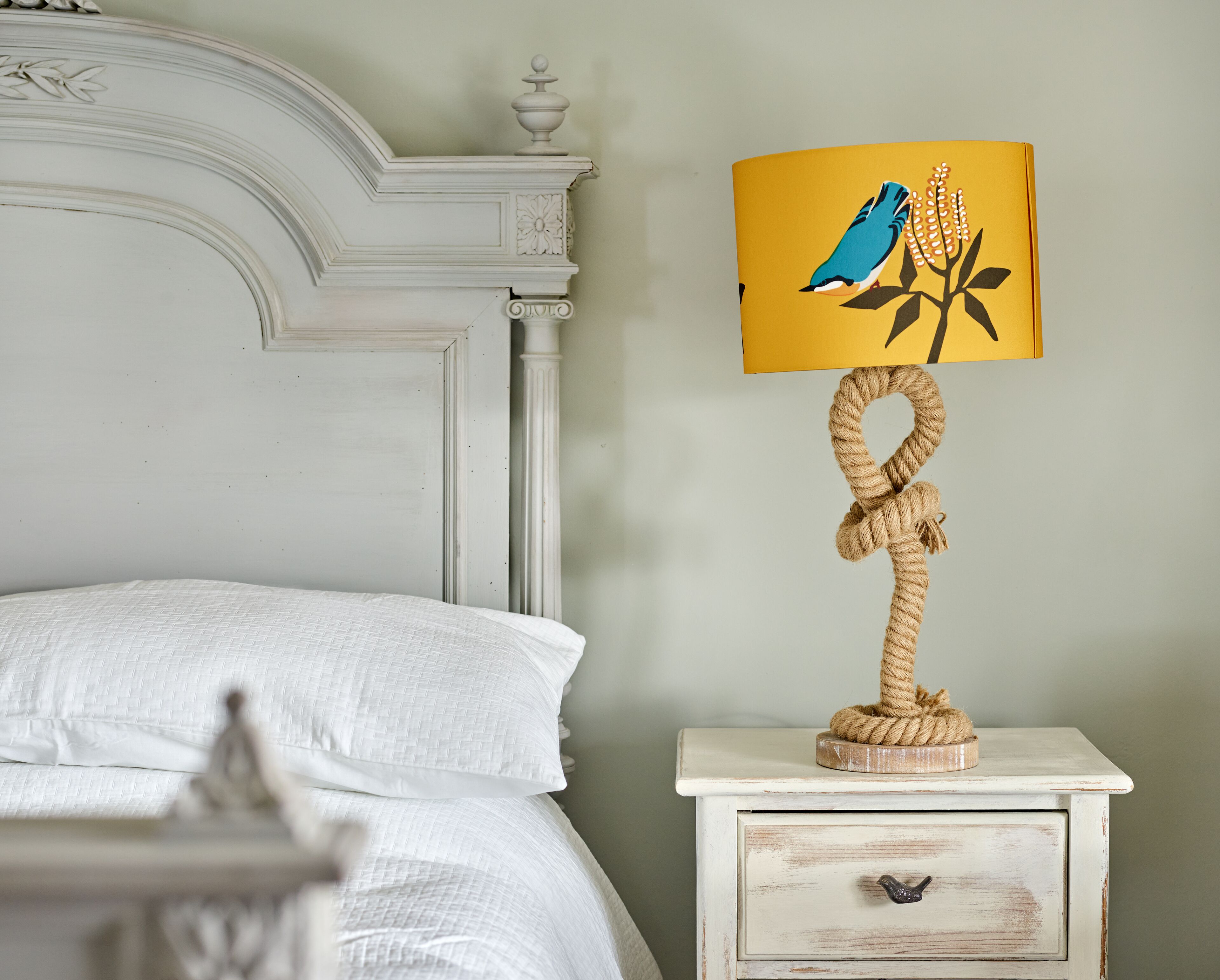 Una cama con ropa de cama blanca, una mesita de noche blanca con lámpara que presenta un diseño de pájaro y planta, y un cajón pequeño.