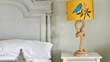 Una cama con ropa de cama blanca, una mesita de noche blanca con lámpara que presenta un diseño de pájaro y planta, y un cajón pequeño.
