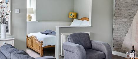 A modern bedroom with a bed, a grey armchair, a wooden dresser, and a small table with glasses and a bottle.