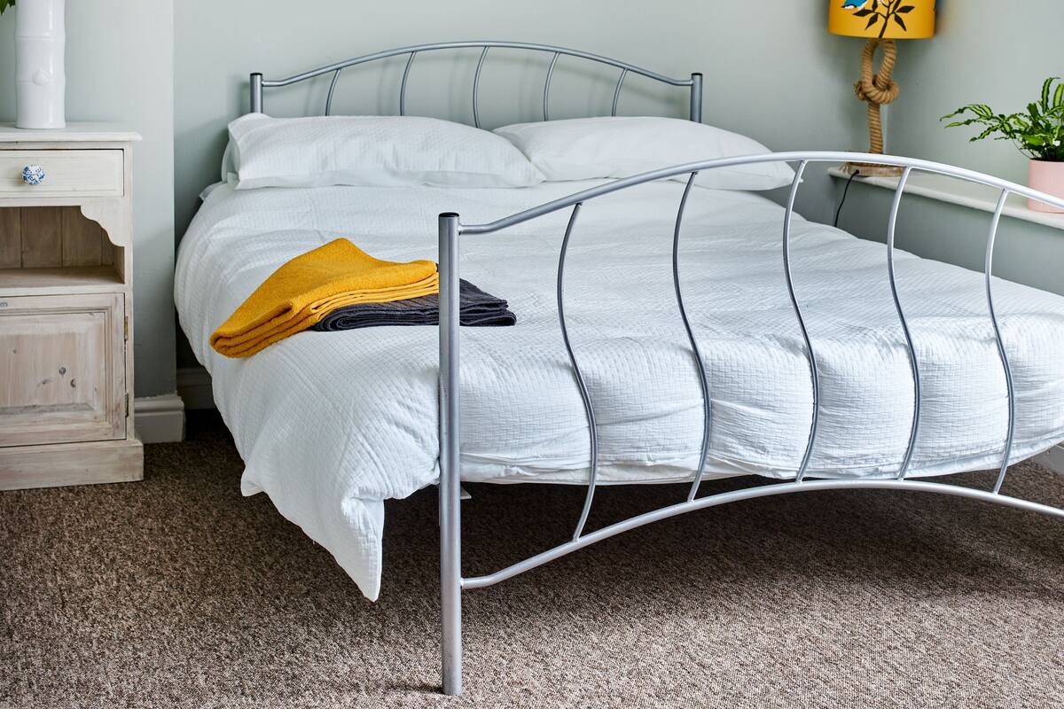 A neatly made bed with a metal frame, a bedside table with a vase of flowers, and a wall-mounted lamp.