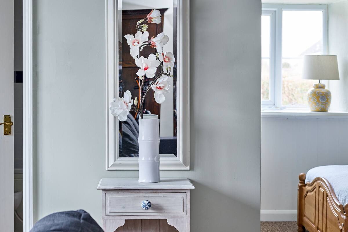 A white bedside table with a vase of white flowers, a wooden nightstand, a bed with a blue blanket, and a window with a lamp on the sill.