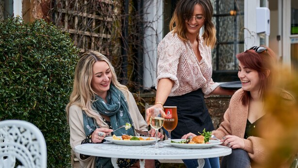 Three people dining at an outdoor table with a white chair, surrounded by greenery and a building in the background. - The Martlet Inn (Wellington)