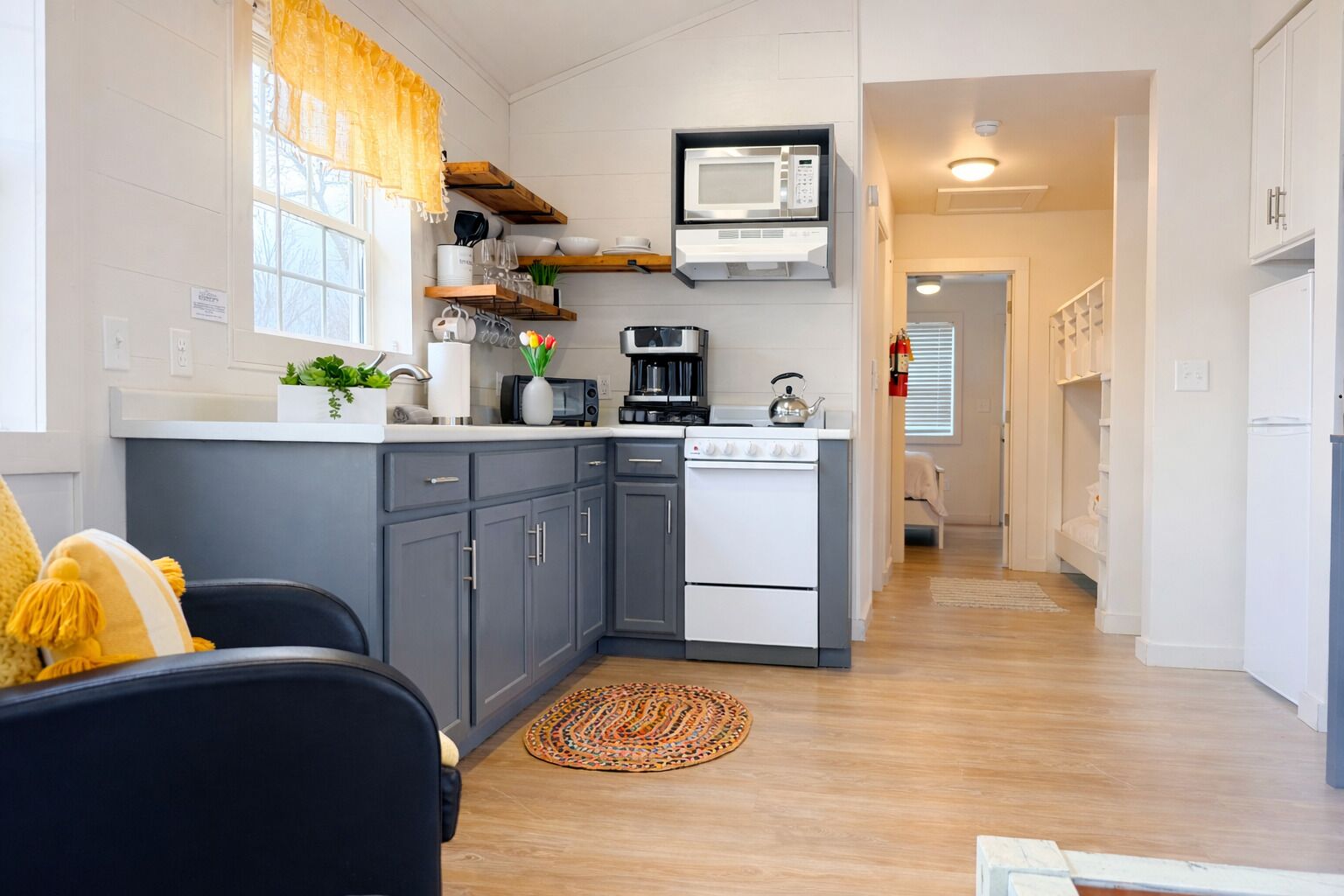 A modern kitchen with grey cabinets, a white stove, and a microwave. There is a window with yellow curtains, a small plant on the counter, and a woven rug on the floor.